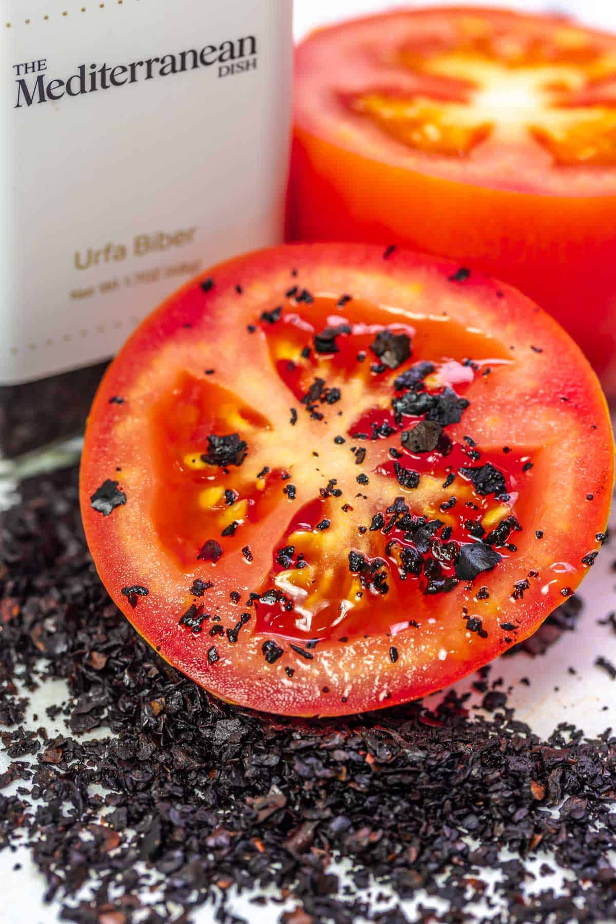 a close up of a bottle of urfa biber spice from The Mediterranean Dish next to two tomato halves sprinkled with the spice.