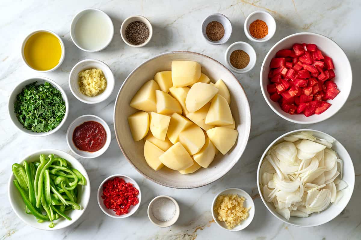 ingredients for dopiazeh aloo perisan potato curry including potatoes, salt, olive oil, onion, bell pepper, garlic, ginger, red chili, turmeric powder, cumin, coriander, tomatoes, tomato paste, lime juice, pepper, and cilantro.