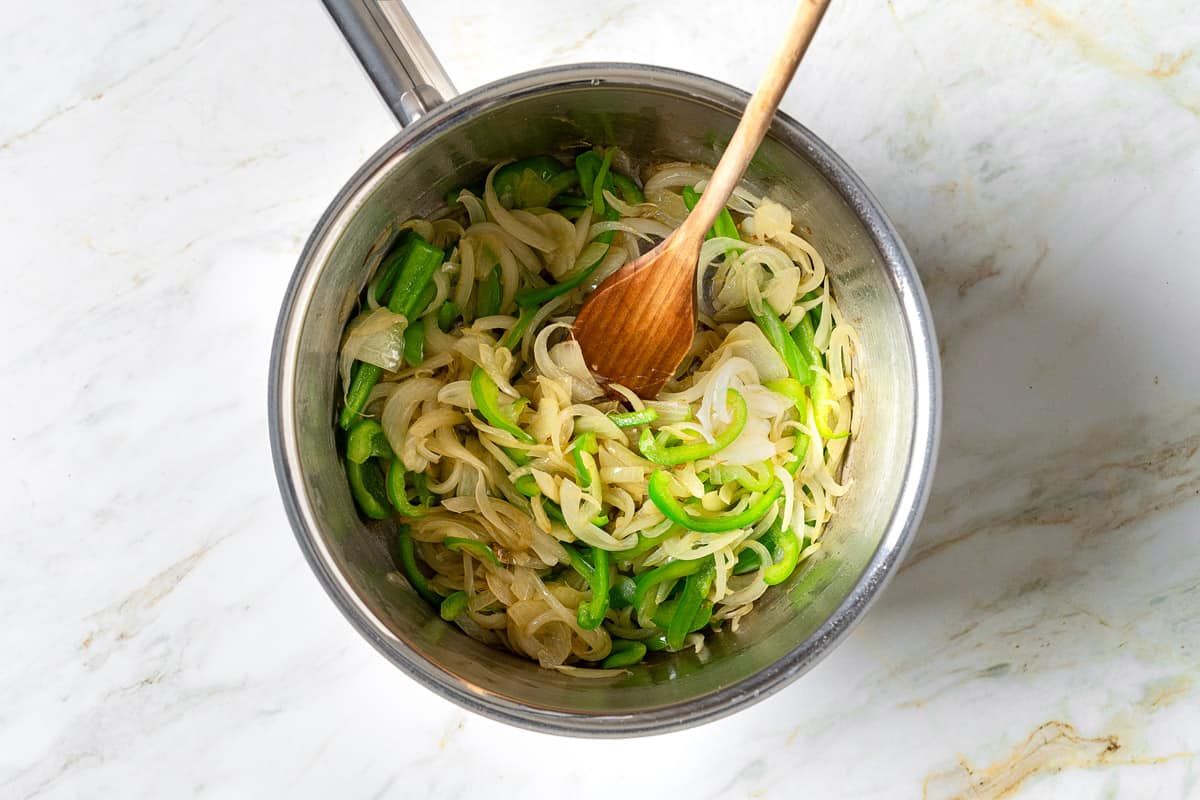 sliced onions and green bell pepper cooking in a saucepan with wooden spoon.