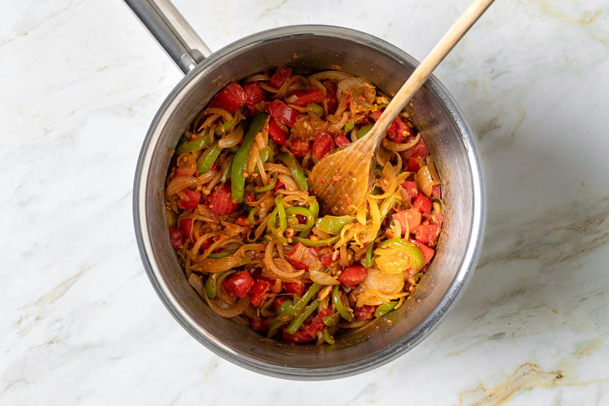 sliced onions, sliced green bell peppers, tomatoes and spices cooking in a saucepan with wooden spoon.