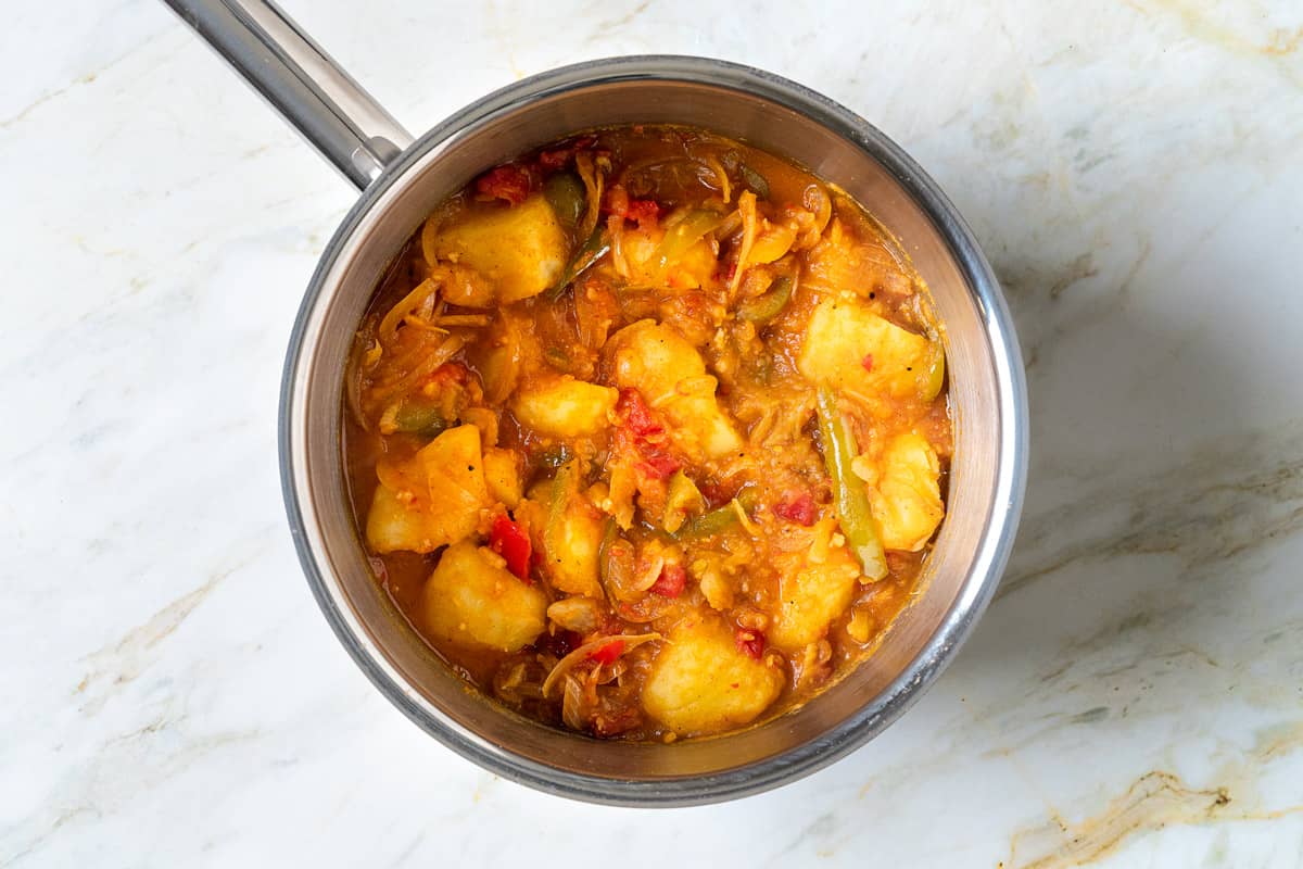 sliced onions, sliced green bell peppers, tomatoes, potatoes and spices cooking in a saucepan.