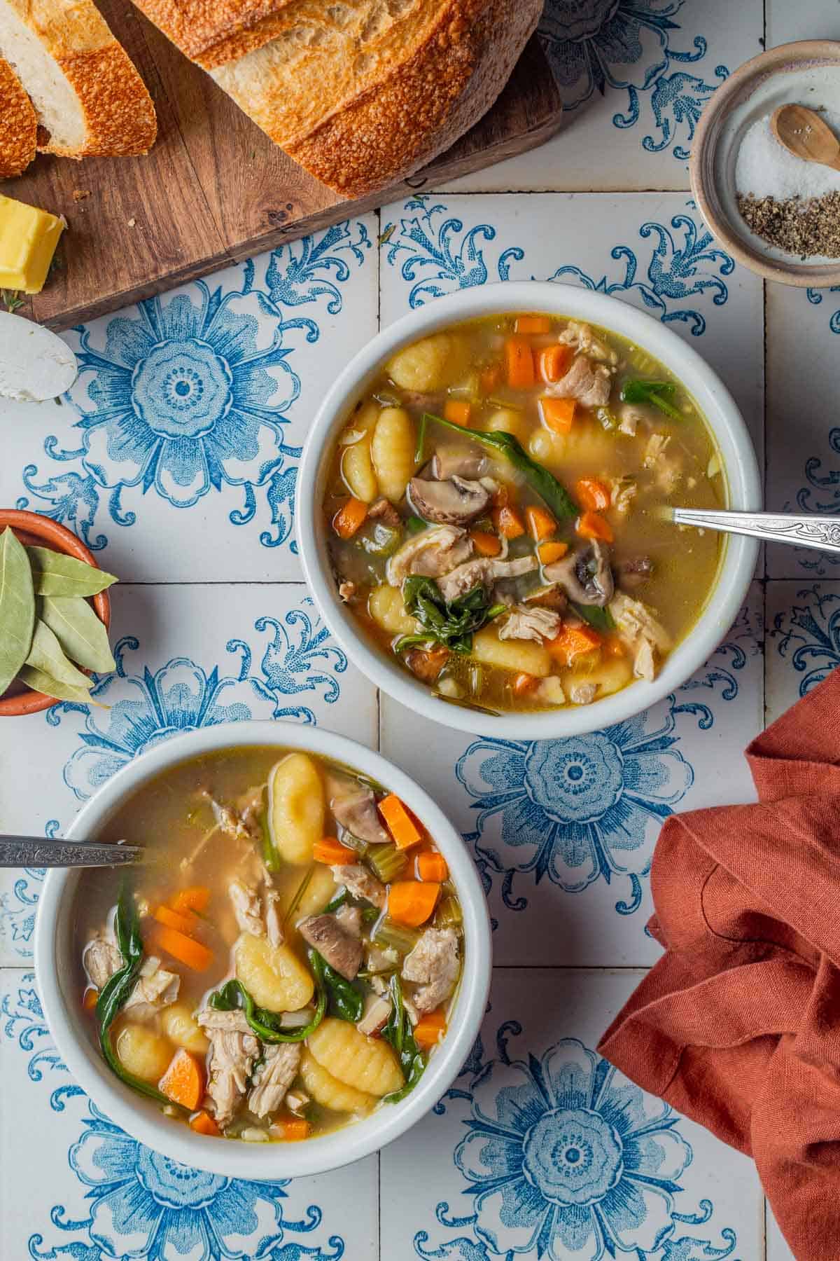 Overhead shot of two bowls of chicken gnocchi soup on white tile with blue flowers and a burnt orange linen on the side.