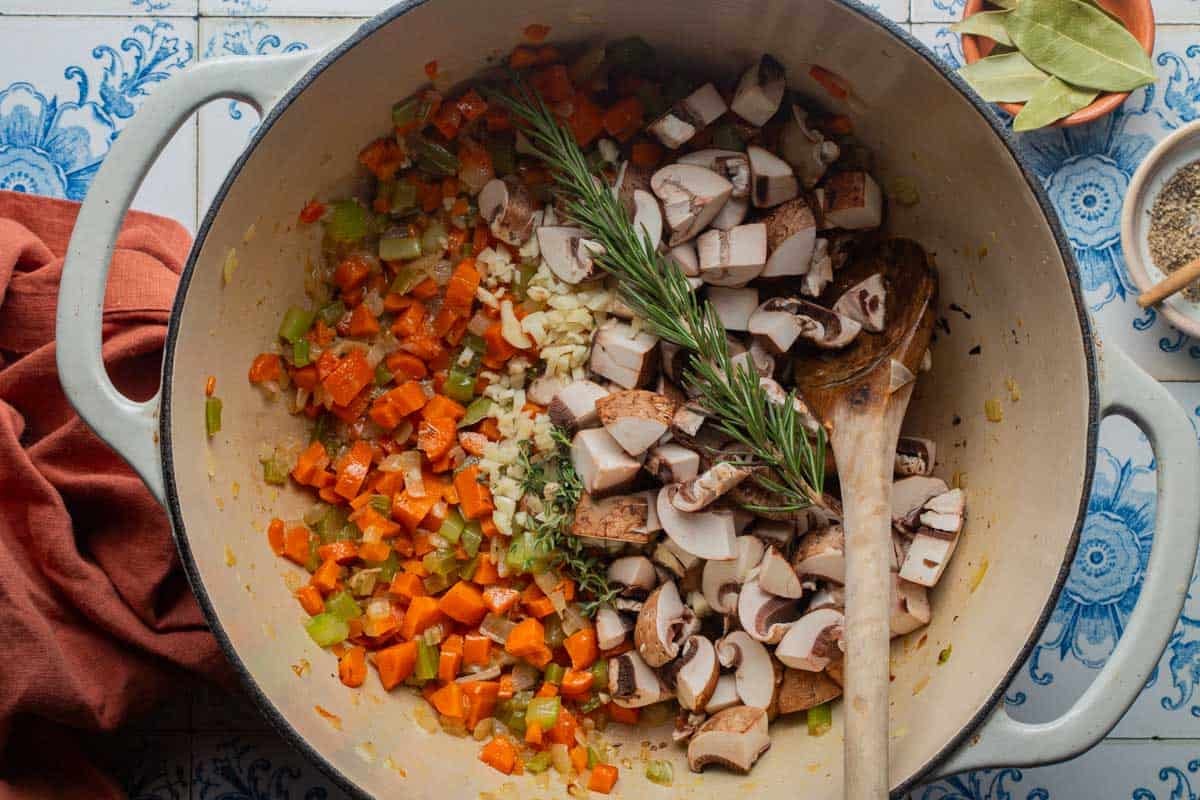 A dutch oven with thyme, rosemary, garlic, and mushrooms that have just been added to the cooked carrot, onion, and celery.