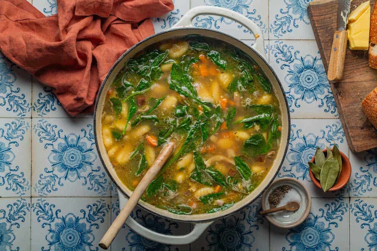 A pot of chicken gnocchi soup with wilted spinach and a wooden spoon.