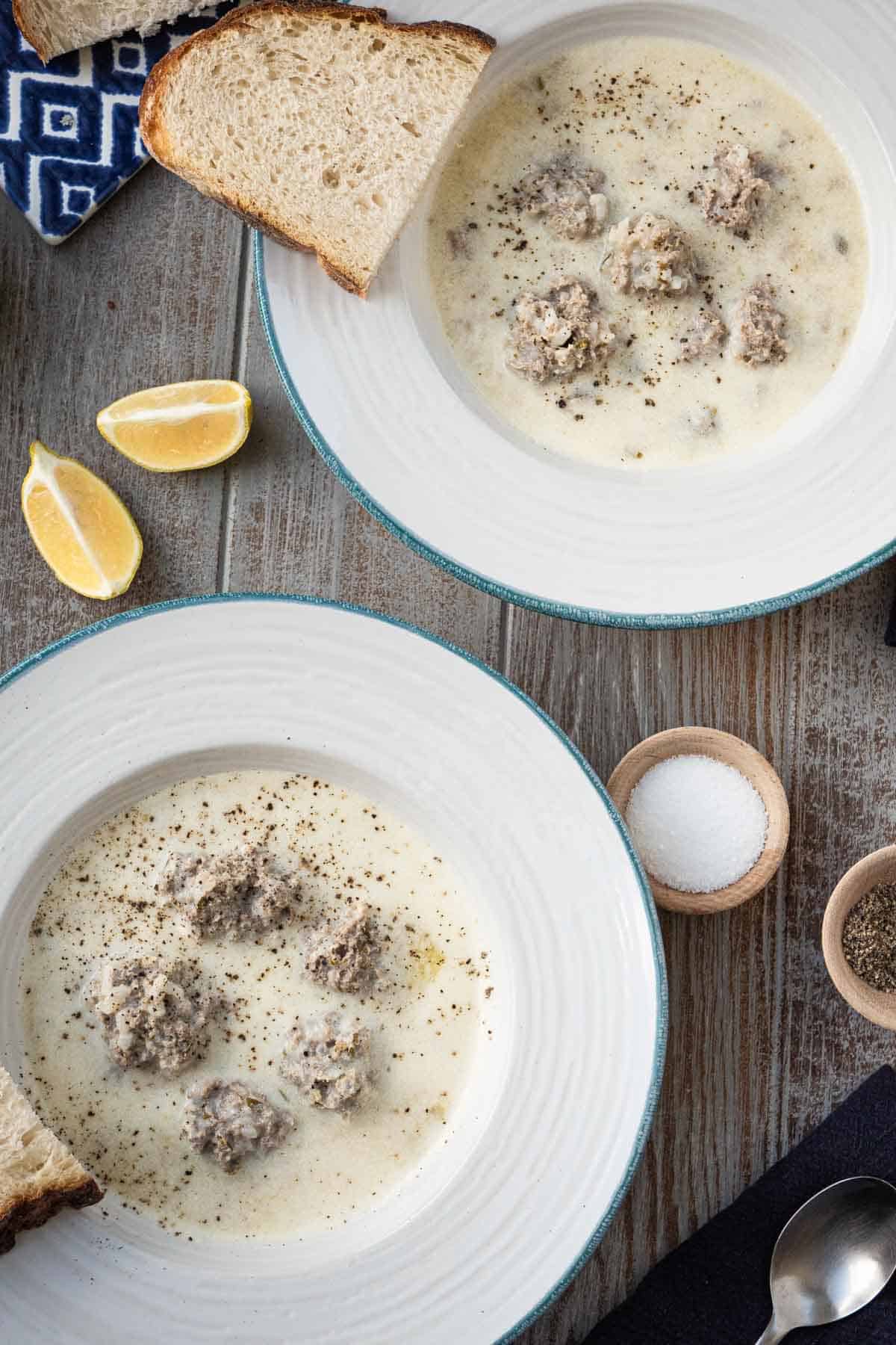 overhead photo of two bowls of Youvarlakia Greek Meatball Soup surrounded by lemon wedges, small bowls of salt and pepper, and a slice of crusty bread.