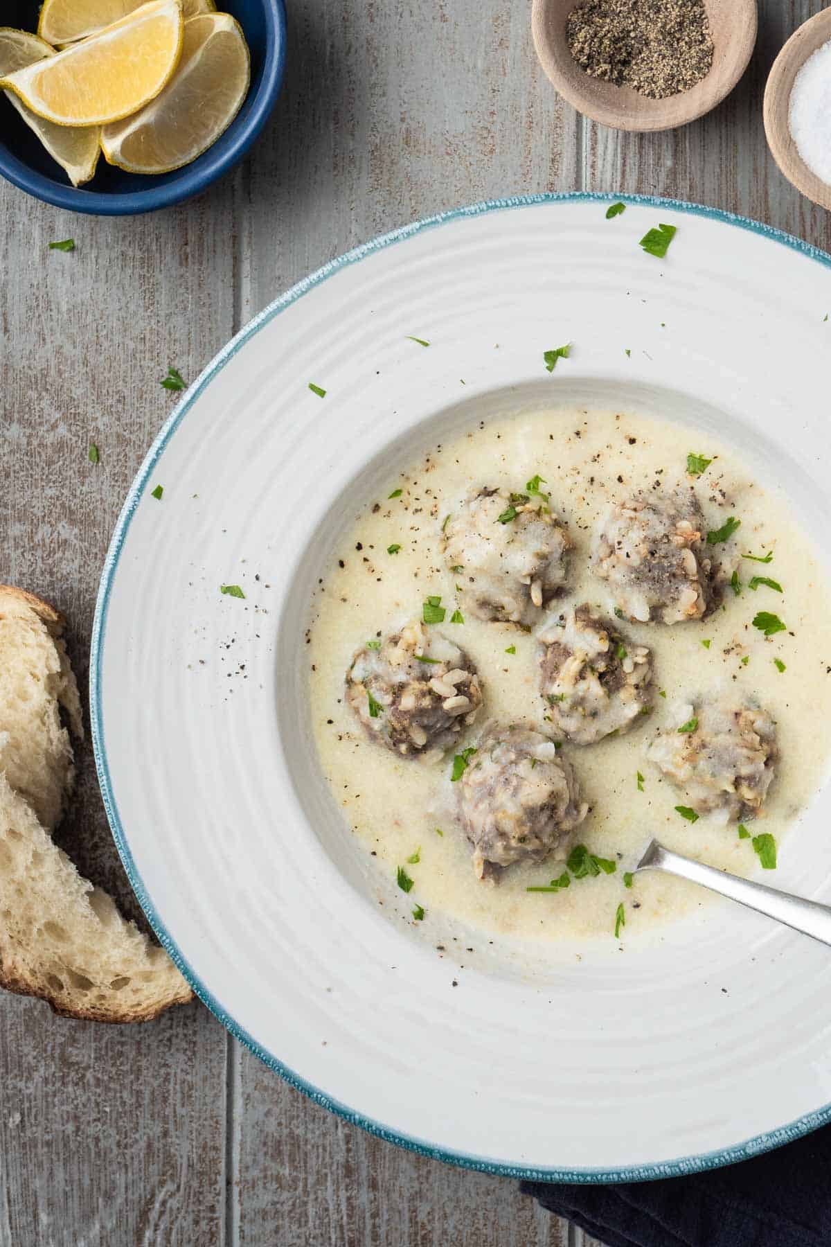 Overhead photo of Youvarlakia Greek Meatball Soup in a bowl surrounded by a small bowl of lemon wedges, bowls of salt and pepper, and a piece of crusty bread.