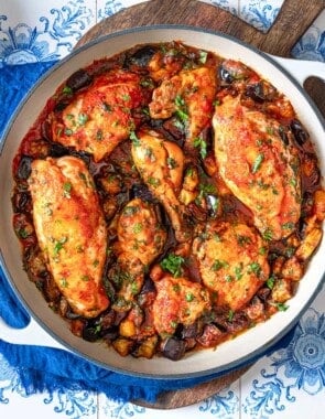 overhead photo of cooked Braised Chicken and Eggplant in a skillet on a wooden serving tray next to a stack of white plates with forks and a small bowl of chopped herbs.