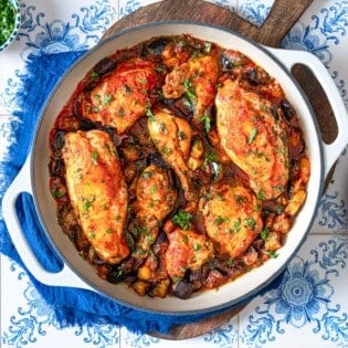 overhead photo of cooked Braised Chicken and Eggplant in a skillet on a wooden serving tray next to a stack of white plates with forks and a small bowl of chopped herbs.