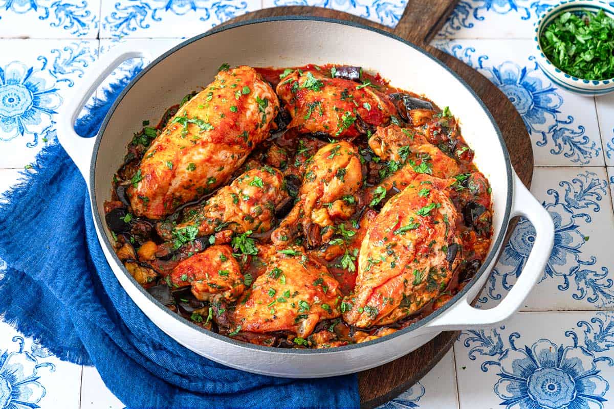 cooked Braised Chicken and Eggplant in a skillet on a wooden serving tray next to a small bowl of chopped herbs.