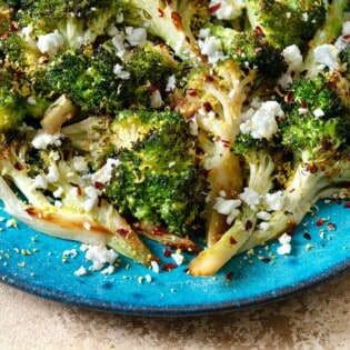 a close up of roasted broccoli topped with feta cheese and red pepper flakes on a blue plate.