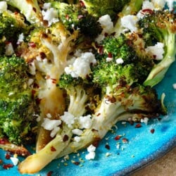 a close up of roasted broccoli topped with feta cheese and red pepper flakes on a blue plate.