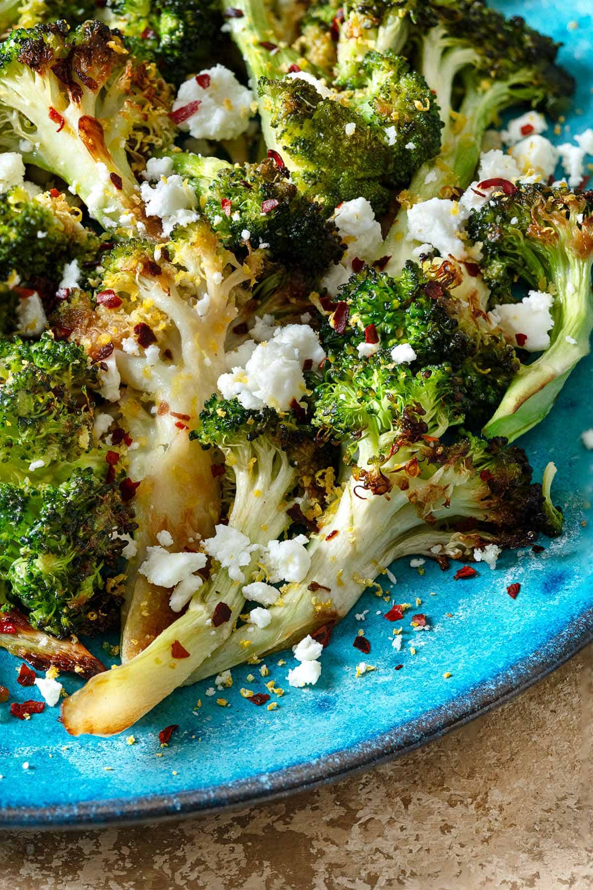 a close up of roasted broccoli topped with feta cheese and red pepper flakes on a blue plate.