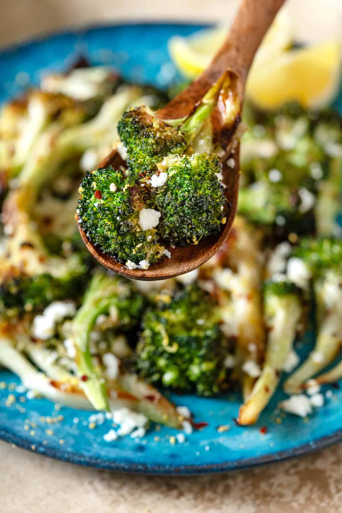 a close up of a bite of roasted broccoli on wooden serving spoon being lifted from the plate.
