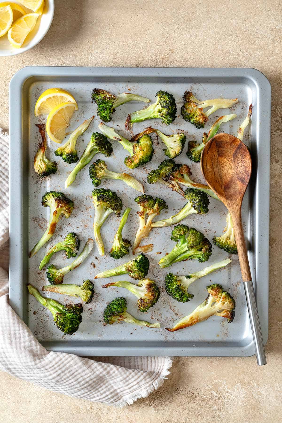 Pieces of roasted broccoli on a sheet pan with 2 lemon wedges and a wooden spoon next to a bowl of lemon wedges.