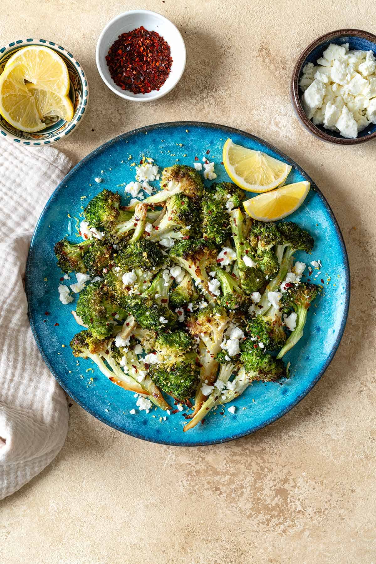 overhead photo of roasted broccoli topped with feta cheese and red pepper flakes with 2 lemon wedges on a blue plate surrounded by of bowls of lemon wedges, red pepper flakes and feta cheese.