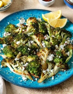 roasted broccoli topped with feta cheese and red pepper flakes with 2 lemon wedges on a blue plate in front of bowls of lemon wedges, red pepper flakes and feta cheese.