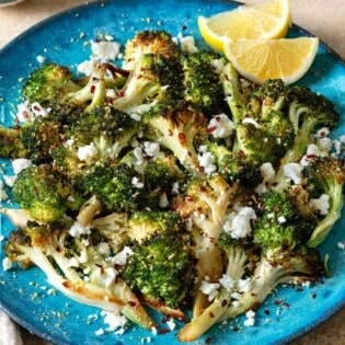 roasted broccoli topped with feta cheese and red pepper flakes with 2 lemon wedges on a blue plate in front of bowls of lemon wedges, red pepper flakes and feta cheese.