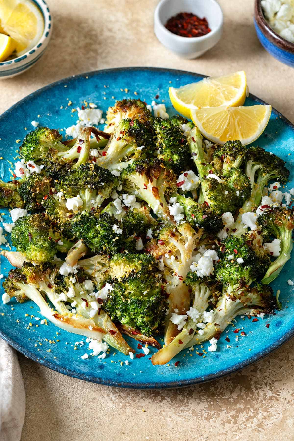 roasted broccoli topped with feta cheese and red pepper flakes with 2 lemon wedges on a blue plate in front of bowls of lemon wedges, red pepper flakes and feta cheese.