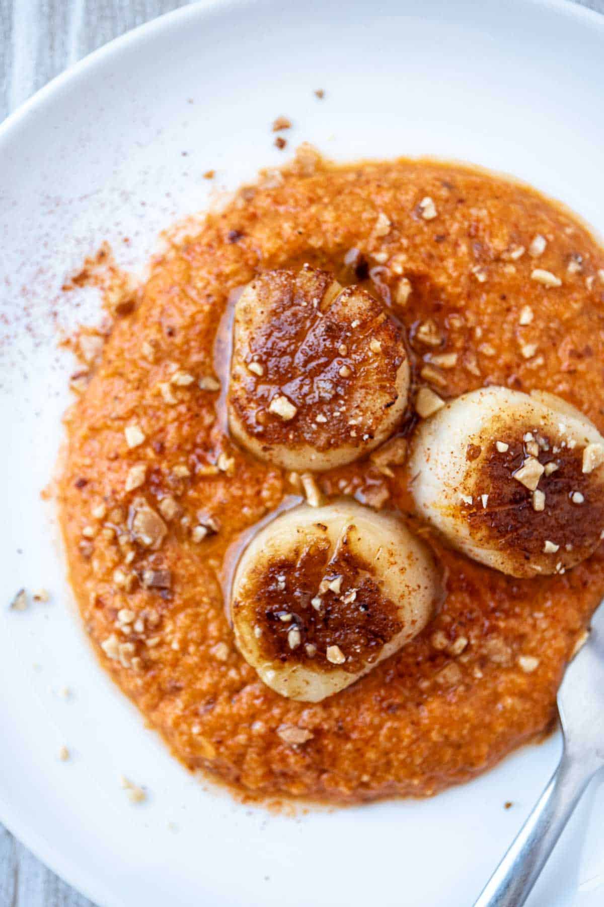 An overhead photo of a plate of seared scallops served on a spicy red pepper sauced and garnished with crushed almonds.