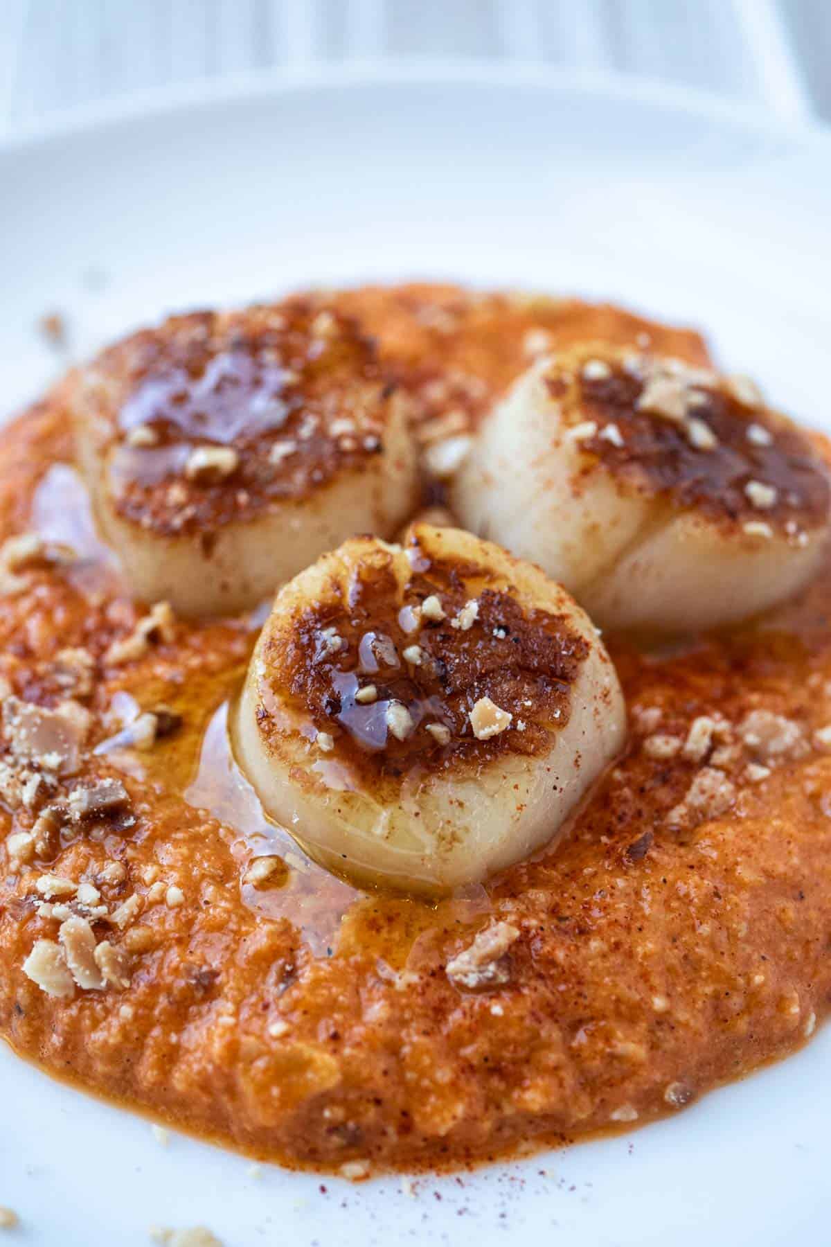 a close up of a plate of seared scallops served on a spicy red pepper sauced and garnished with crushed almonds.