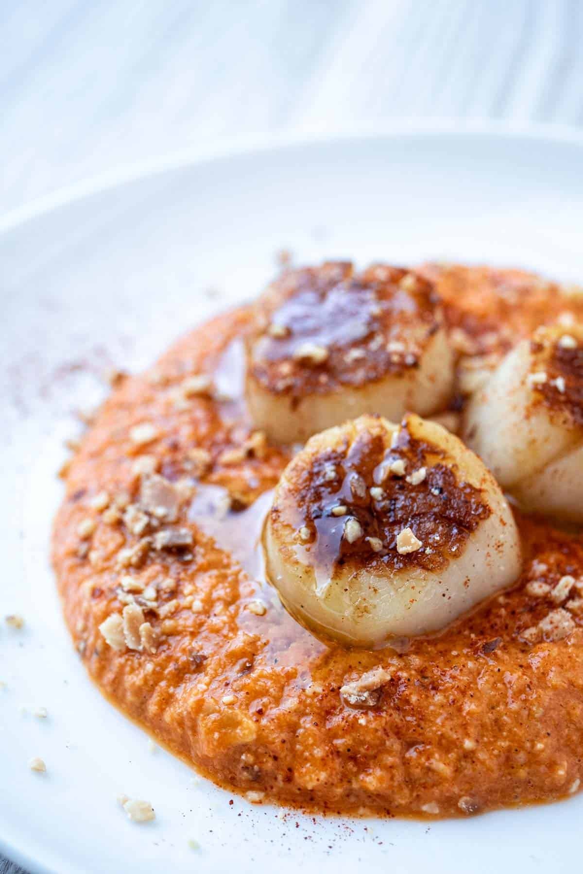 a close up of a plate of seared scallops served on a spicy red pepper sauced and garnished with crushed almonds.