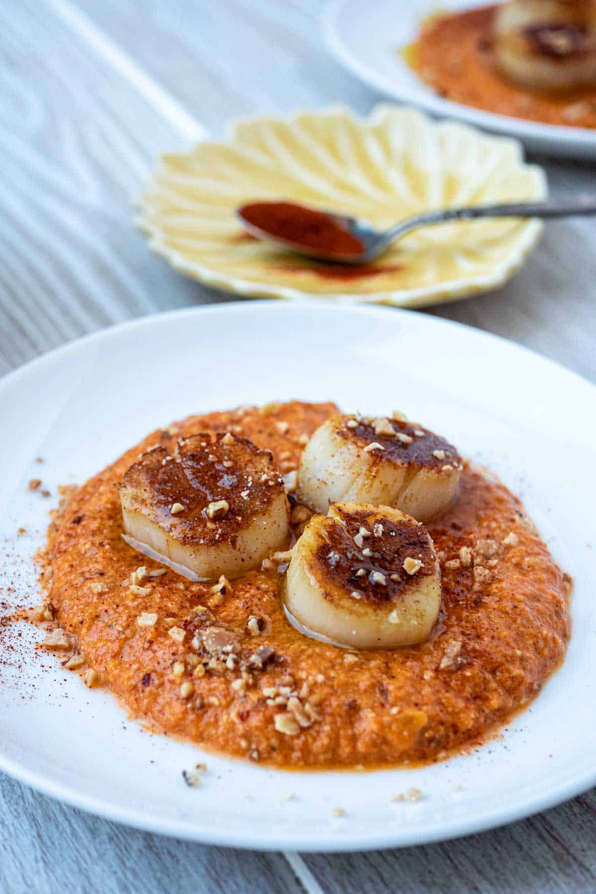 a plate of seared scallops served on a spicy red pepper sauced and garnished with crushed almonds in front of a small plate with a spoon of paprika.