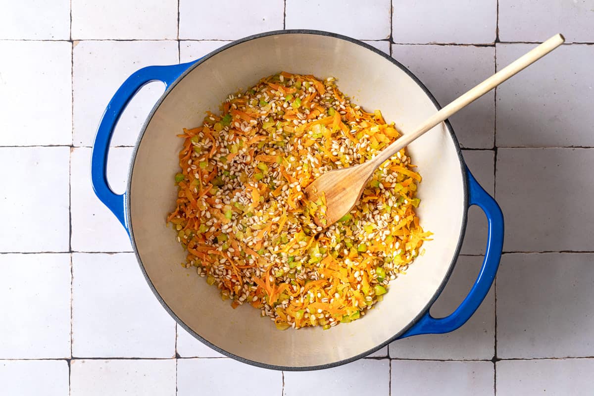 vegetables and barley being sautéed in a pot with a wooden spoon.