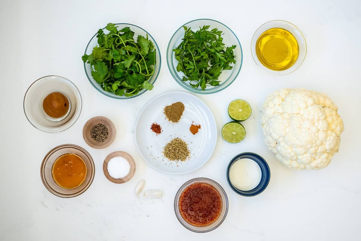 Ingredients for cauliflower steak, including olive oil, one head of cauliflower, harissa paste, garlic, honey, dried oregano, fresh lime, Greek yogurt, Salt, and pepper.