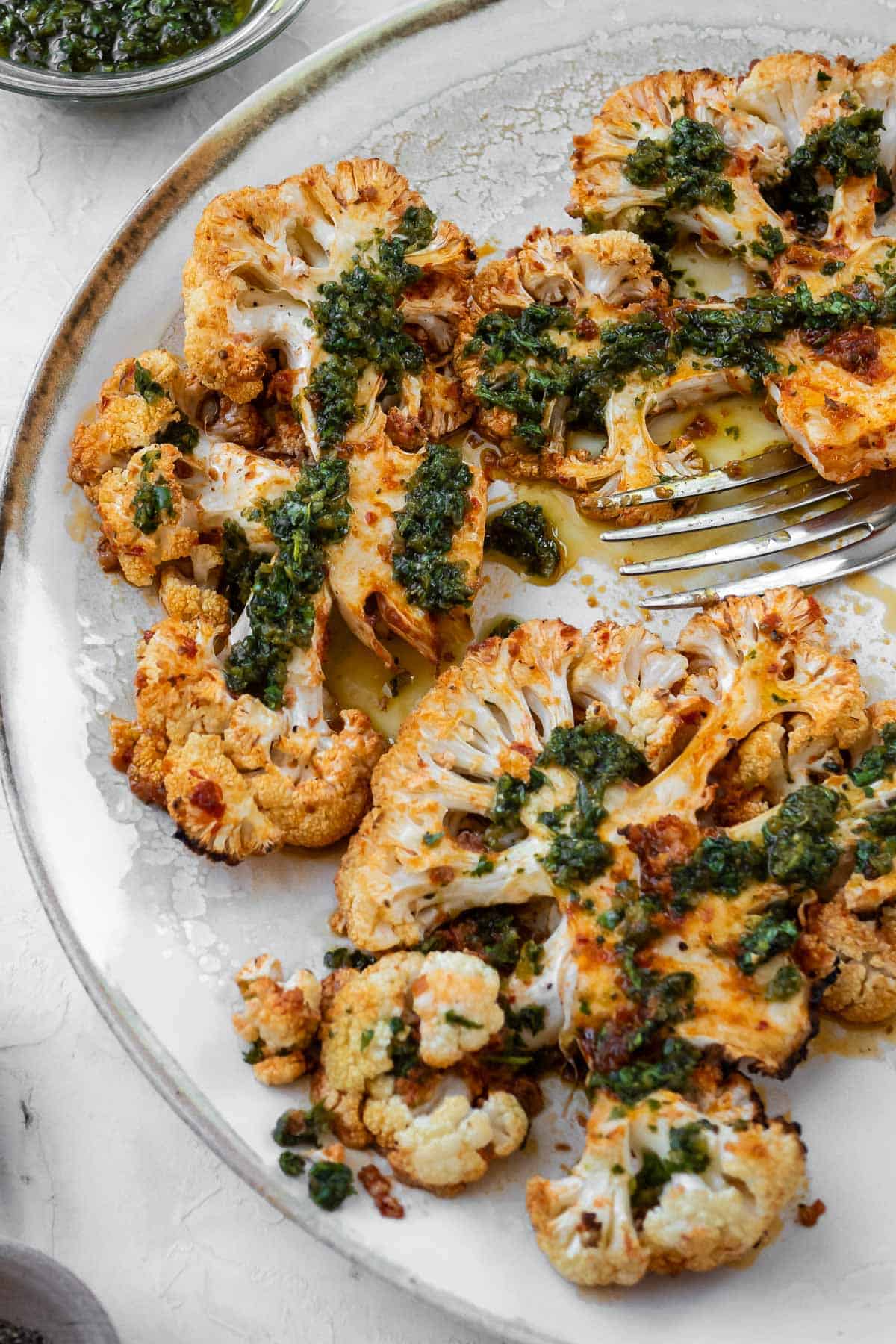 Cauliflower steaks on a white serving plate finished with Chermoula with a small bowl of Chermoula on the side.