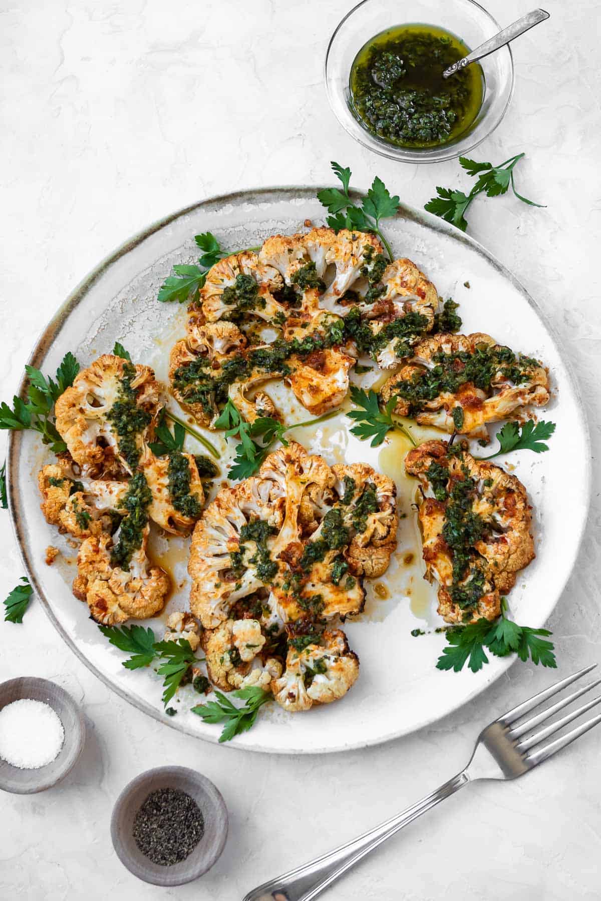 Overhead shot of golden brown cauliflower steaks on a white serving plate. The steaks were finished with Chermoula and fresh parsley leaves, and there is a small bowl of Chermoula on the side.