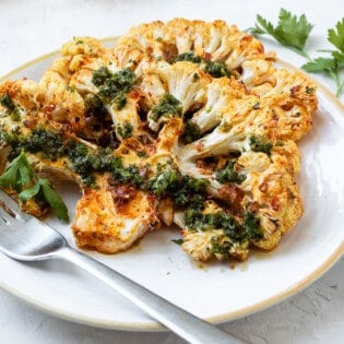 Side shot of a cauliflower steak on a white plate with a metal fork.