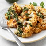 Two cauliflower steaks attractively layered on a plate with one resting on top of the other. Both have been dressed with green Chermoula. There is a small bowl of Chermoula on the side and a metal fork on the plate.