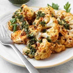 Two cauliflower steaks attractively layered on a plate with one resting on top of the other. Both have been dressed with green Chermoula. There is a small bowl of Chermoula on the side and a metal fork on the plate.