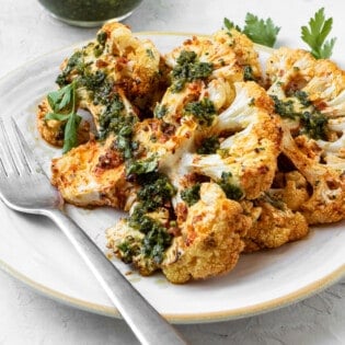 Two cauliflower steaks attractively layered on a plate with one resting on top of the other. Both have been dressed with green Chermoula. There is a small bowl of Chermoula on the side and a metal fork on the plate.