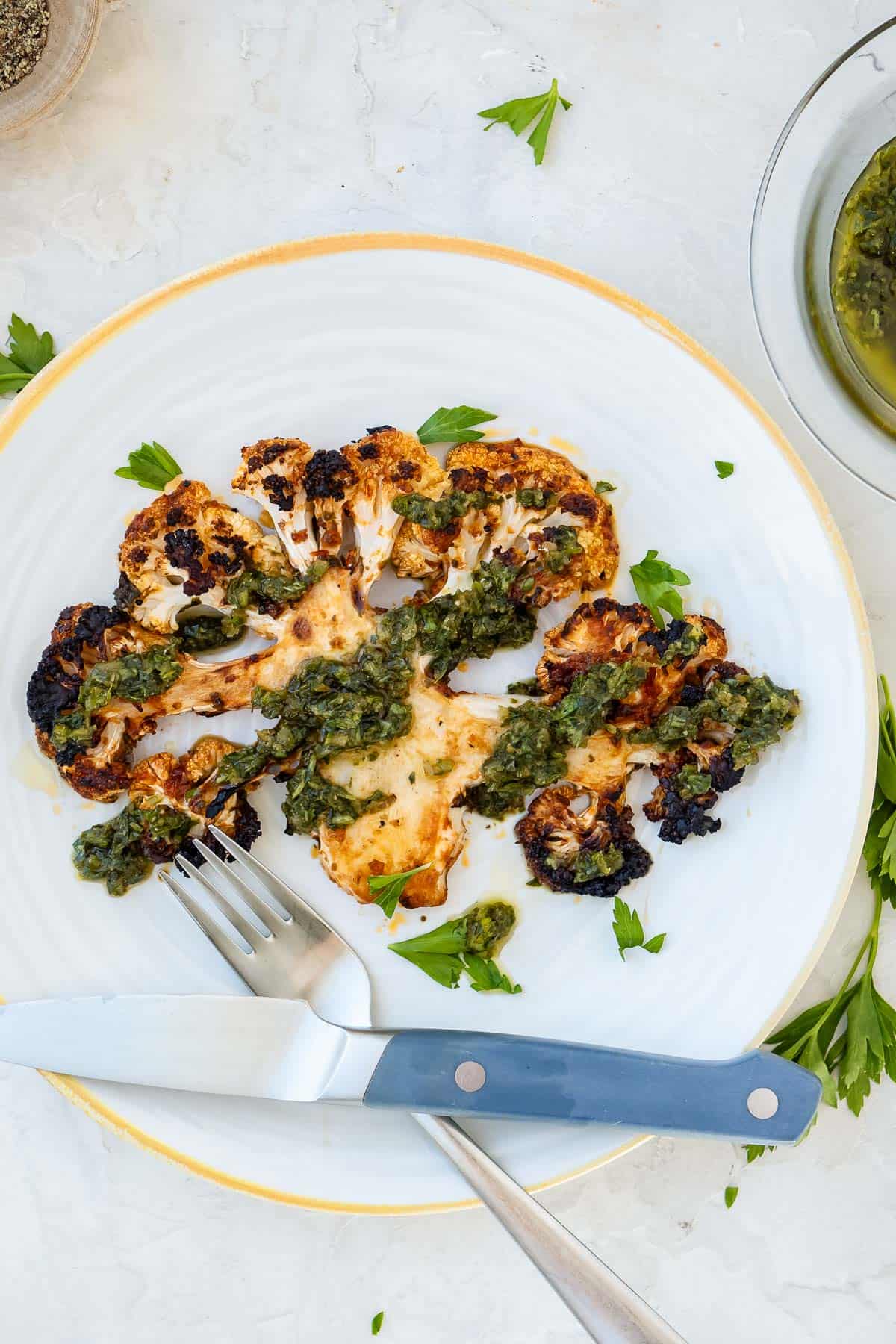 Overhead shot of cauliflower steak on a white plate with a fork and knife crossing on the plate and chermoula dressing on top.