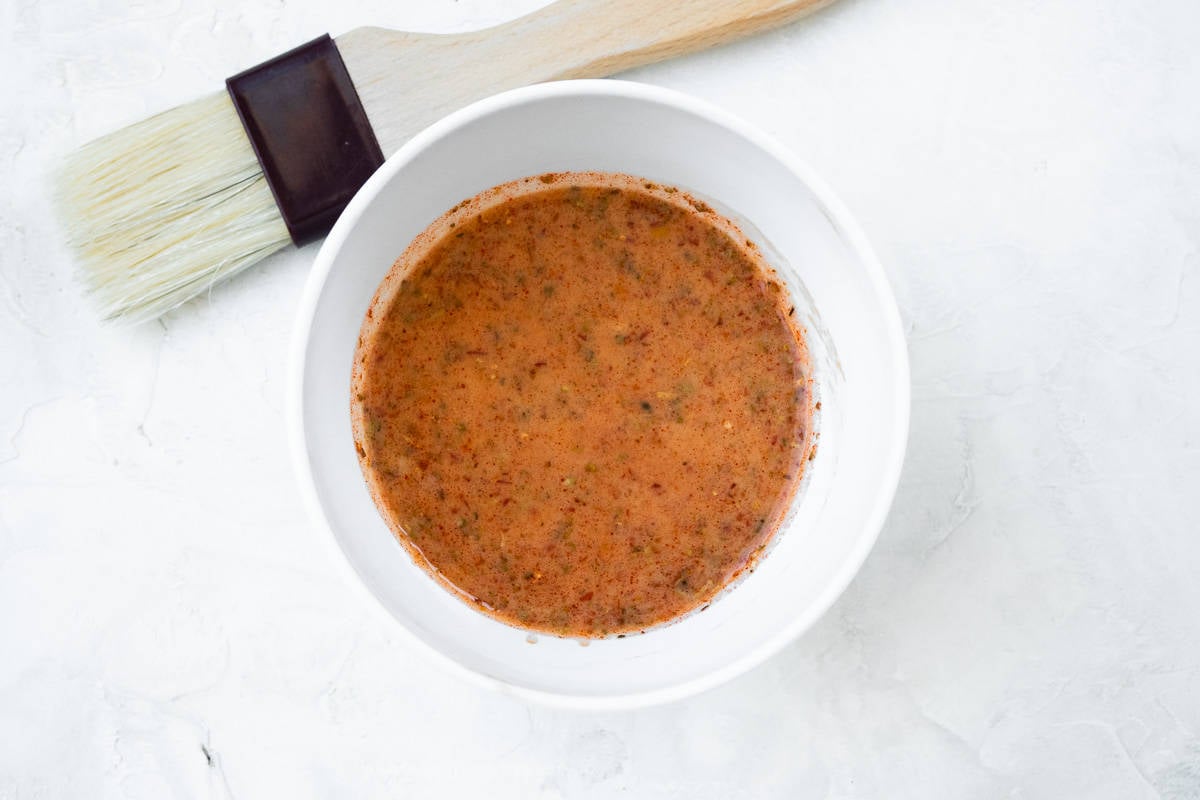 A white bowl showing a red harissa glaze with flecks of oregano and a pastry brush on the side.