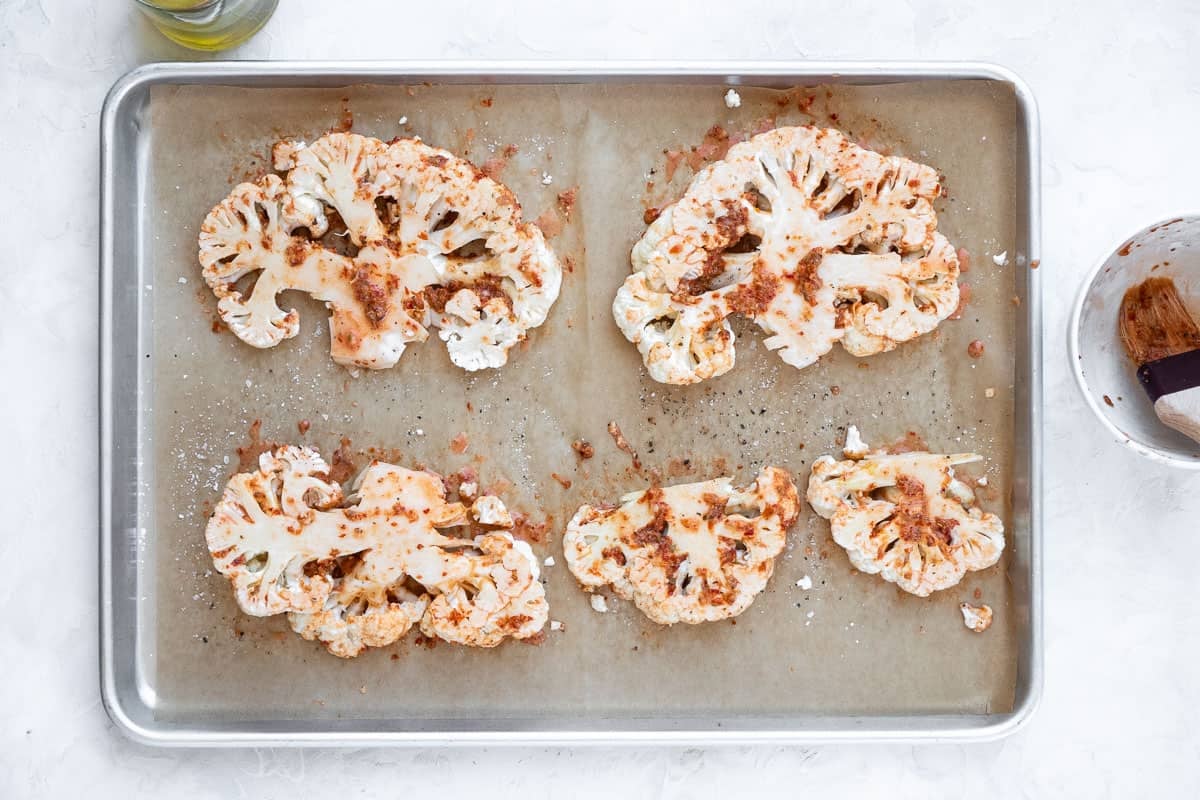 Five cauliflower planks on a parchment-lined sheet tray. The cauliflower planks have been seasoned with salt and pepper and brushed with harissa glaze. They are spaced apart to allow them to crisp and turn golden brown.