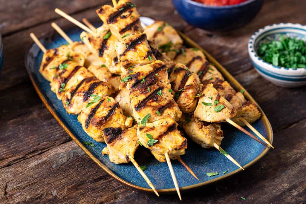 a close up of 6 joojeh kabobs on a serving platter next to a small bowl of parsley.