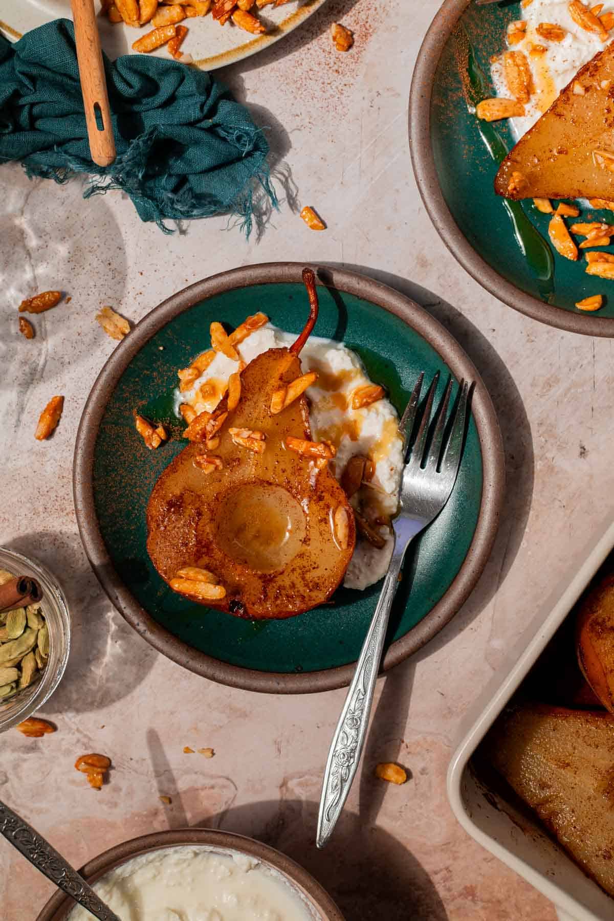 Overhead shot of a serving of baked pears. There is a large spoonful of ricotta cheese, a baked pear resting with its cut side facing up, and candied almonds sprinkled over top. It's all finished with a drizzle of honey.