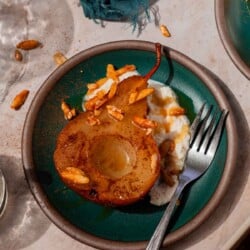 Teal plate with a large spoonful of ricotta cheese, a baked pear resting with its cut side facing up, and candied almonds sprinkled over top. It's all finished with a drizzle of honey.