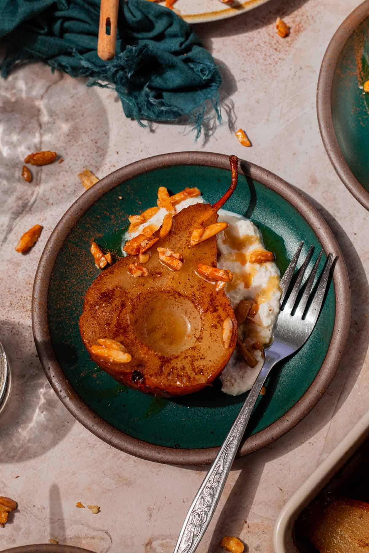 Teal plate with a large spoonful of ricotta cheese, a baked pear resting with its cut side facing up, and candied almonds sprinkled over top. It's all finished with a drizzle of honey.