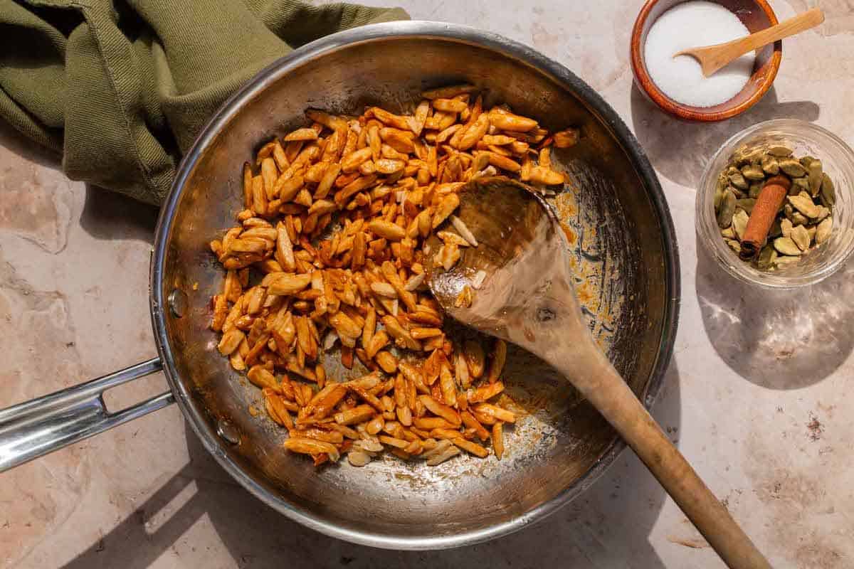 A saucepan with almonds coated in honey, olive oil, and salt being stirred with a wooden spoon.