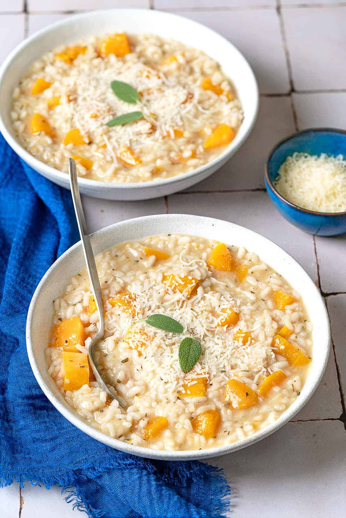 two bowls of butternut squash risotto, one with a spoon, next to a small bowl of parmigiano-reggiano cheese.