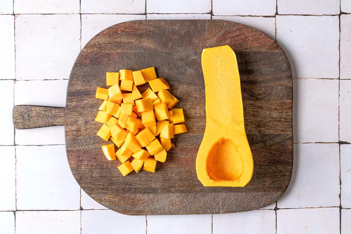 a butternut squash half next to a pile of cubed butternut squash on a wooden cutting board.