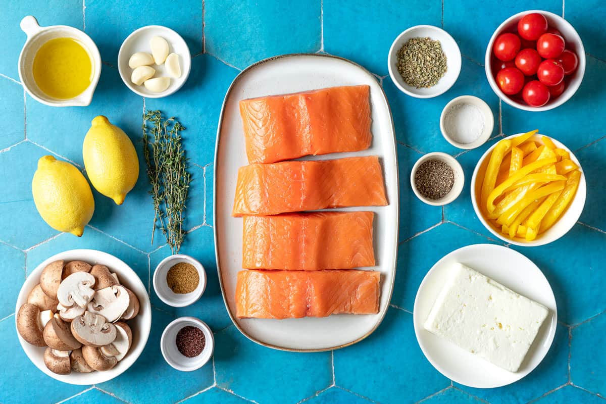 ingredients for easy baked salmon with vegetables and feta including salmon fillets, feta, cherry tomatoes, bell pepper, mushrooms, lemons, garlic, oregano, sumac, cumi, garlic, thyme, salt, pepper, and olive oil.