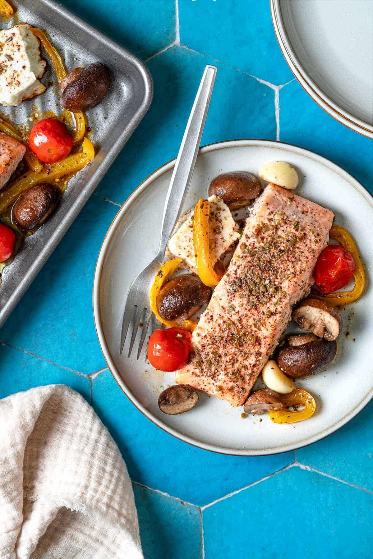 overhead photo of a baked salmon fillet with veggies and feta on a white plate with a fork nest to a sheet pan with baked veggie and feta and a stack of plates.