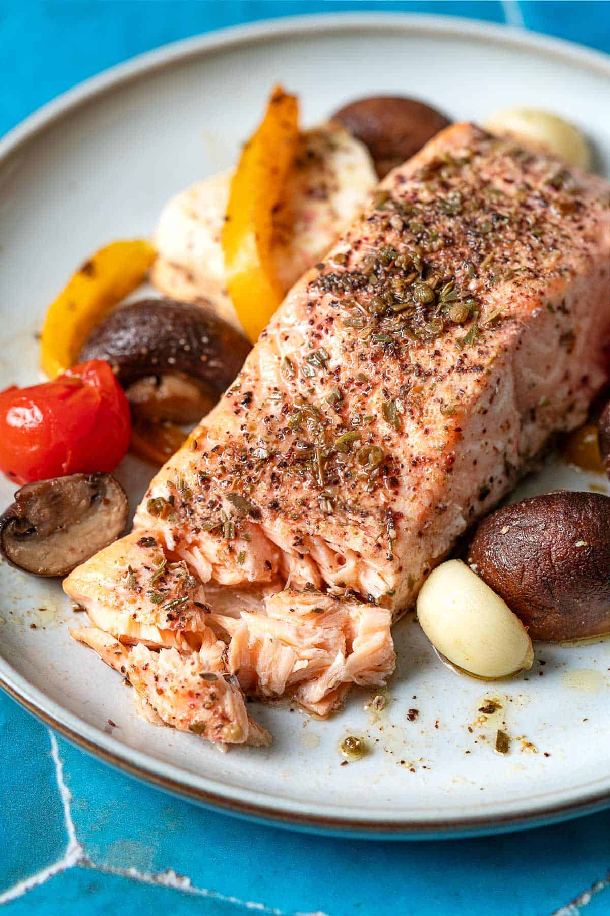 a close up photo of a baked salmon fillet with veggies and feta on a white plate.