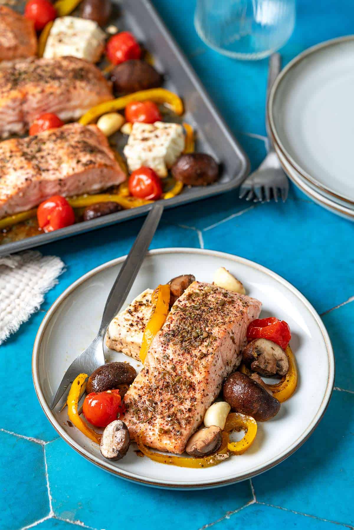 a baked salmon fillet with veggies and feta on a white plate with a fork in front of a sheet pan with baked veggie and feta, a glass, a stack of plates, and 2 forks.