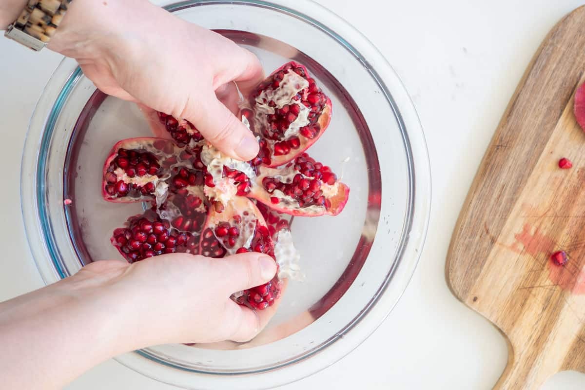 two hands pulling apart sections of a pomegranate and adding each section to the bowl of cold water.