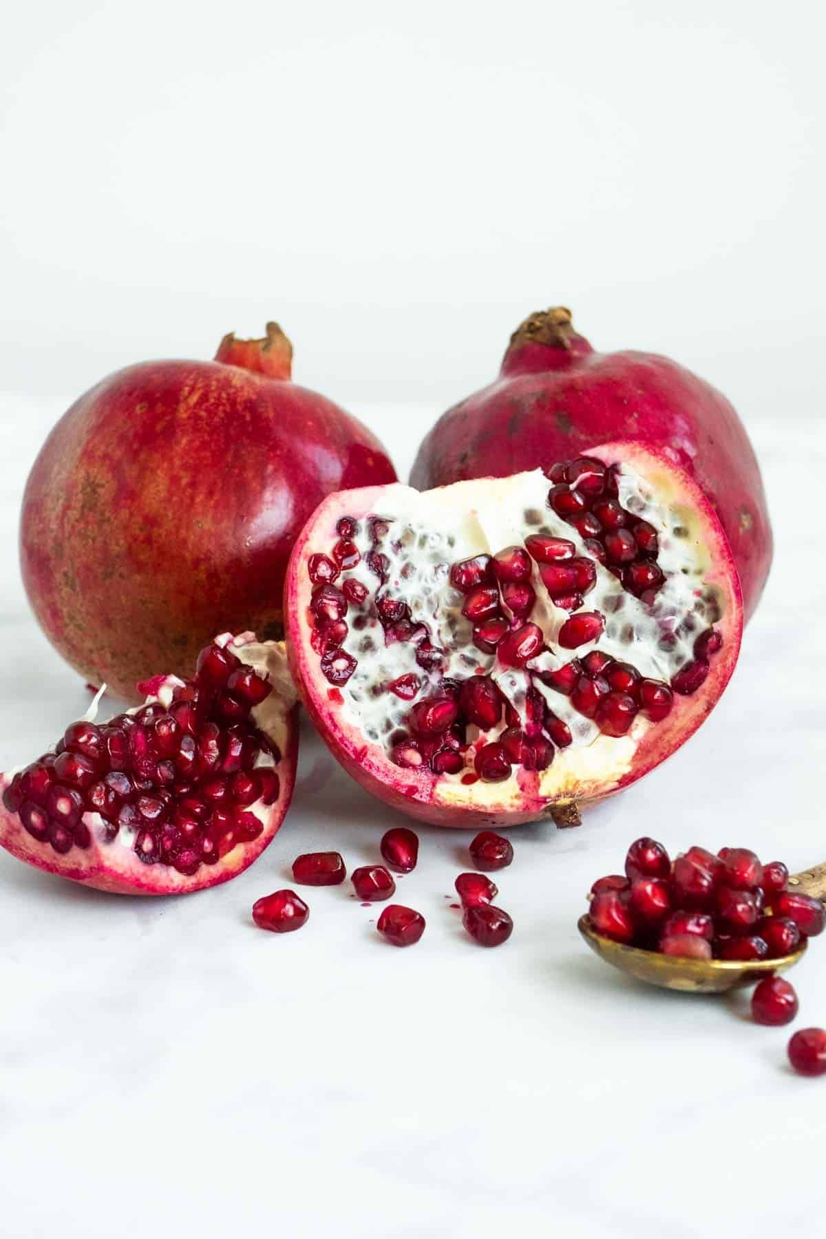a close up of 3 pomegranates, two whole and one sectioned exposing the inside next to a gold spoon containing pomegranate seeds.